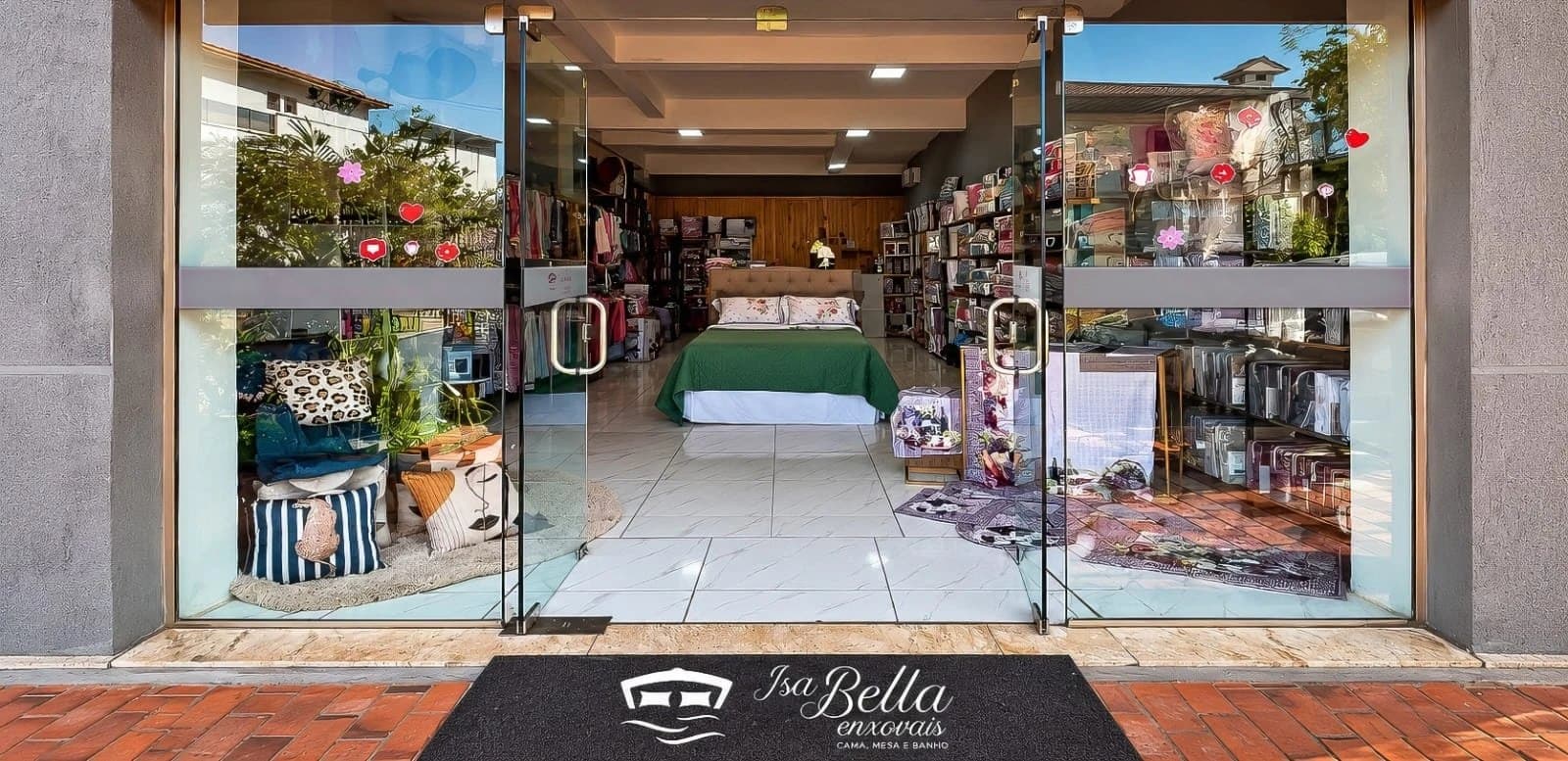 Fachada da Isabella Enxovais com as portas de vidro abertas: almofadas e tapetes na vitrine, uma cama arrumada e prateleiras de enxovais lá dentro e, na entrada, o tapete com o logo da loja