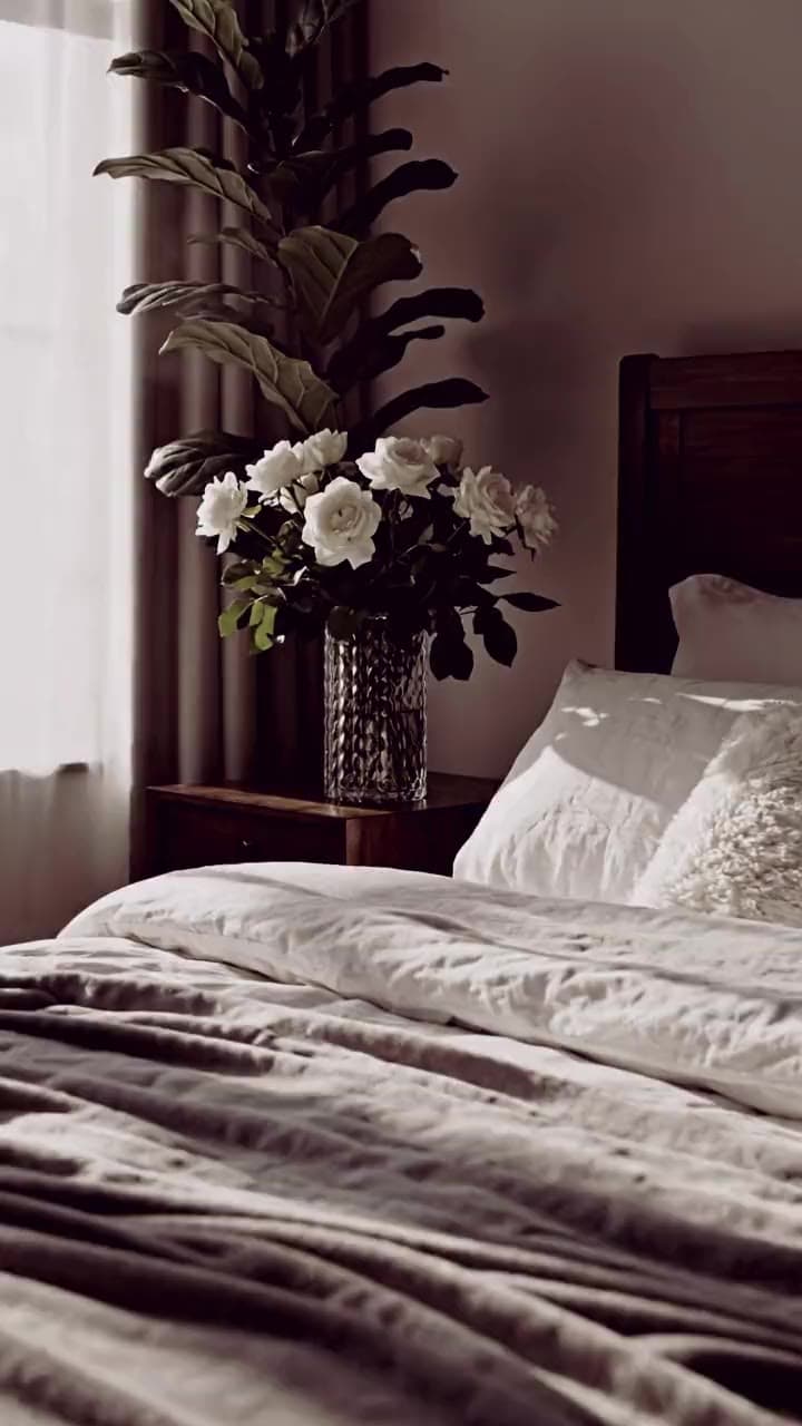 Quarto com a cama arrumada em roupa de cama clara, uma planta e a luz da janela